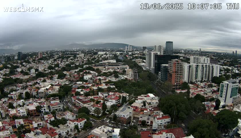 An aerial view of a sprawling city under a cloudy sky shows a mix of residential neighborhoods and taller buildings, with mountains visible in the distance.