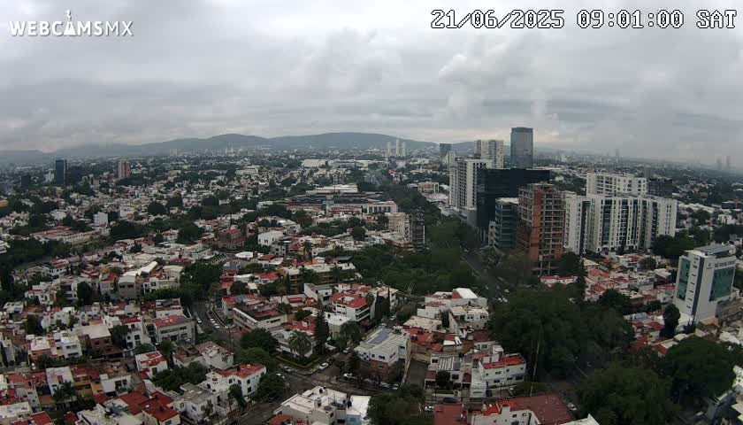 An aerial view of a city on an overcast day, showing a mix of residential buildings and taller structures, with mountains visible in the distance.