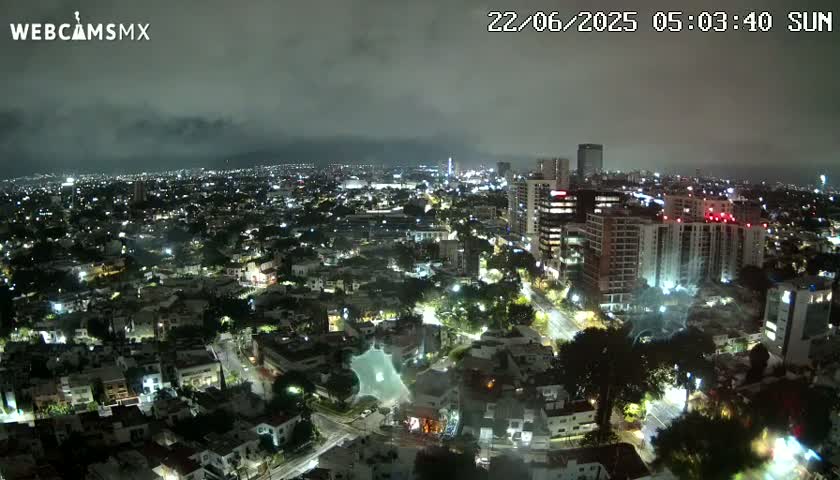 A nighttime aerial view of a city with overcast skies shows a sprawling cityscape illuminated by streetlights and building lights.