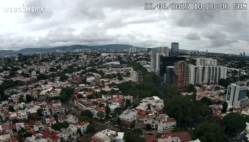 An aerial view of a sprawling city on a cloudy day shows many residential buildings interspersed with taller structures, mountains in the distance, and abundant green spaces between the buildings.