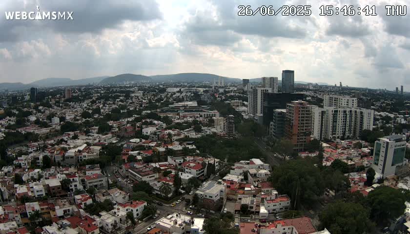 A sprawling city landscape under a partly cloudy sky, with numerous residential buildings and taller structures, extending to distant mountains.