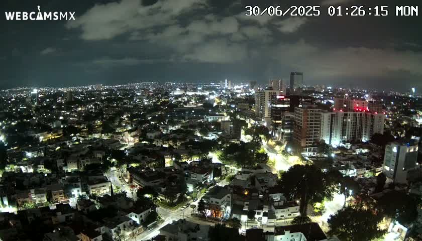 A nighttime aerial view of a city under a partly cloudy sky shows many illuminated buildings and streets.