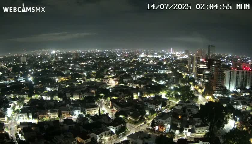 A nighttime aerial view of a sprawling city with many illuminated buildings and streets under a partly cloudy sky.