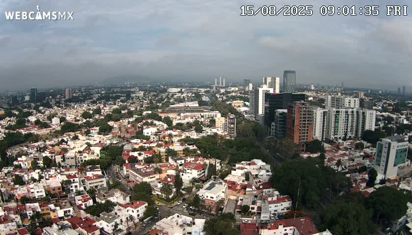 An aerial view shows a sprawling city under a partly cloudy sky, with mountains visible in the distance.