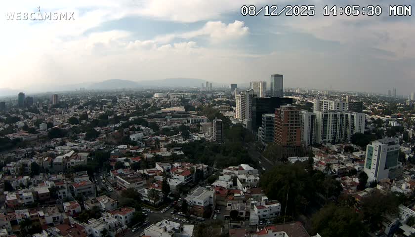 An expansive daytime aerial view reveals a sprawling city featuring a mix of low-rise residential areas and numerous modern high-rise buildings, all under a hazy sky with scattered clouds.