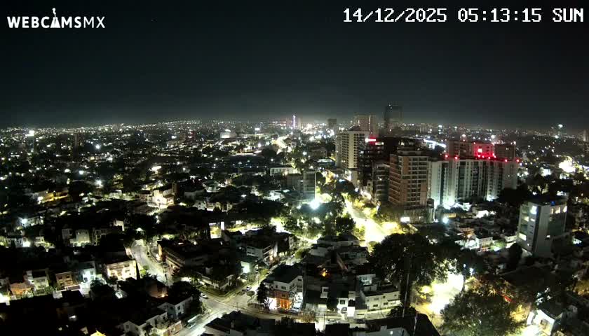 An expansive daytime aerial view reveals a sprawling city featuring a mix of low-rise residential areas and numerous modern high-rise buildings, all under a hazy sky with scattered clouds.