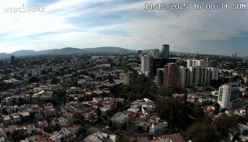 An expansive daytime aerial view reveals a sprawling city featuring a mix of low-rise residential areas and numerous modern high-rise buildings, all under a hazy sky with scattered clouds.