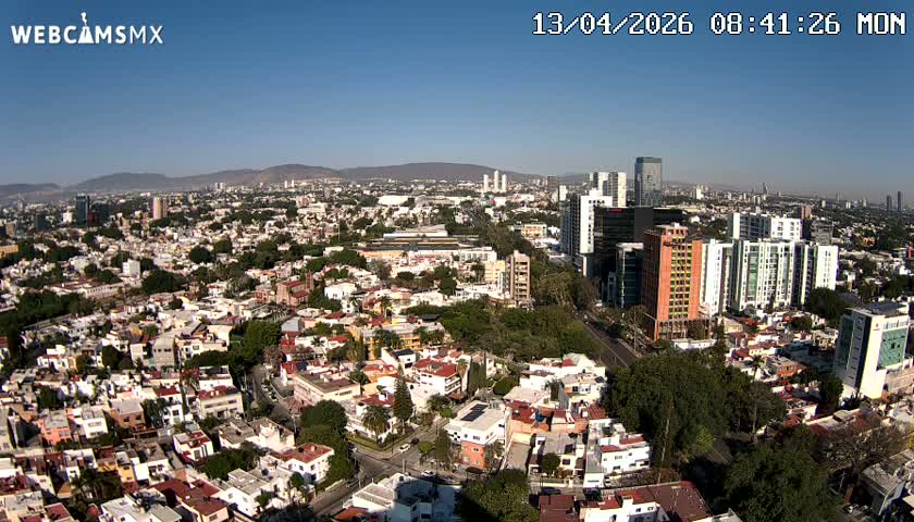 An expansive daytime aerial view reveals a sprawling city featuring a mix of low-rise residential areas and numerous modern high-rise buildings, all under a hazy sky with scattered clouds.
