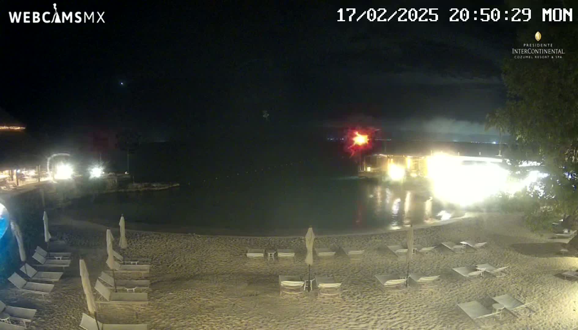 A nighttime beach scene shows a sandy shore with empty lounge chairs and umbrellas, a lit pool, and a dark body of water beyond.