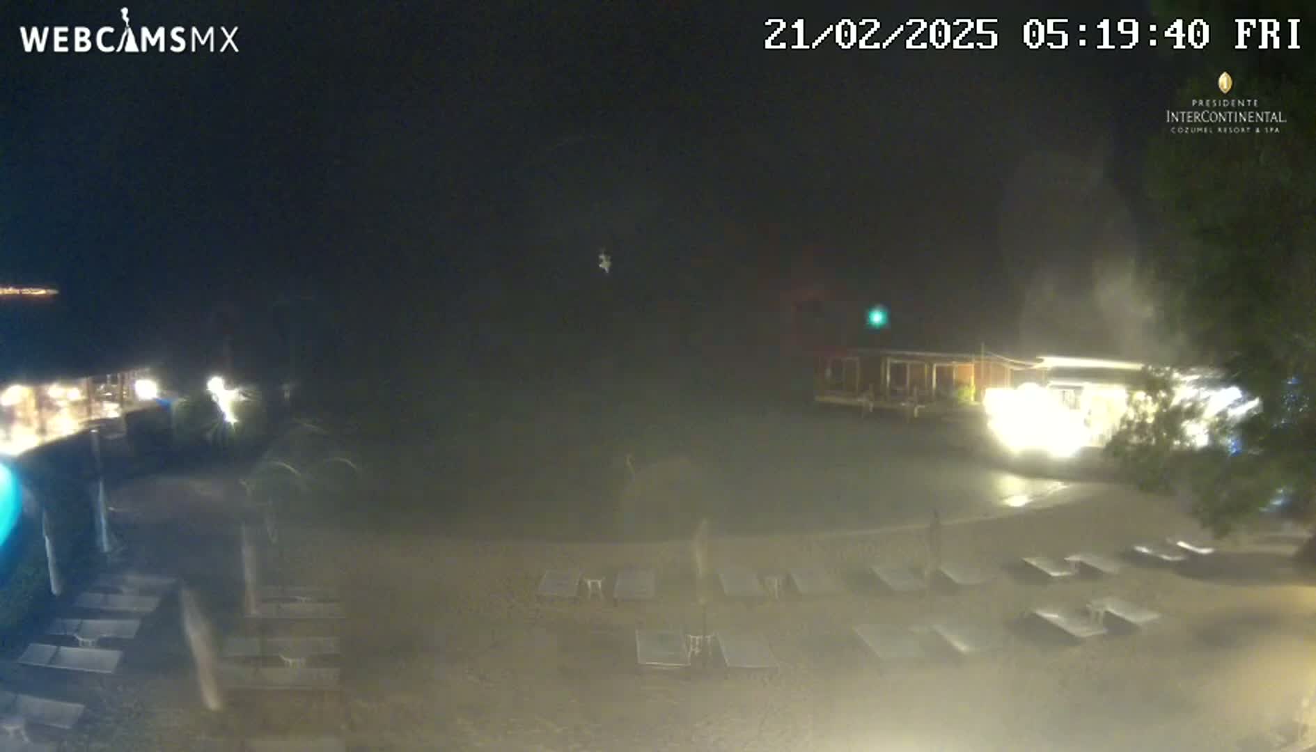 A hazy night scene shows a beach with empty lounge chairs, a building with lights, and some palm trees.