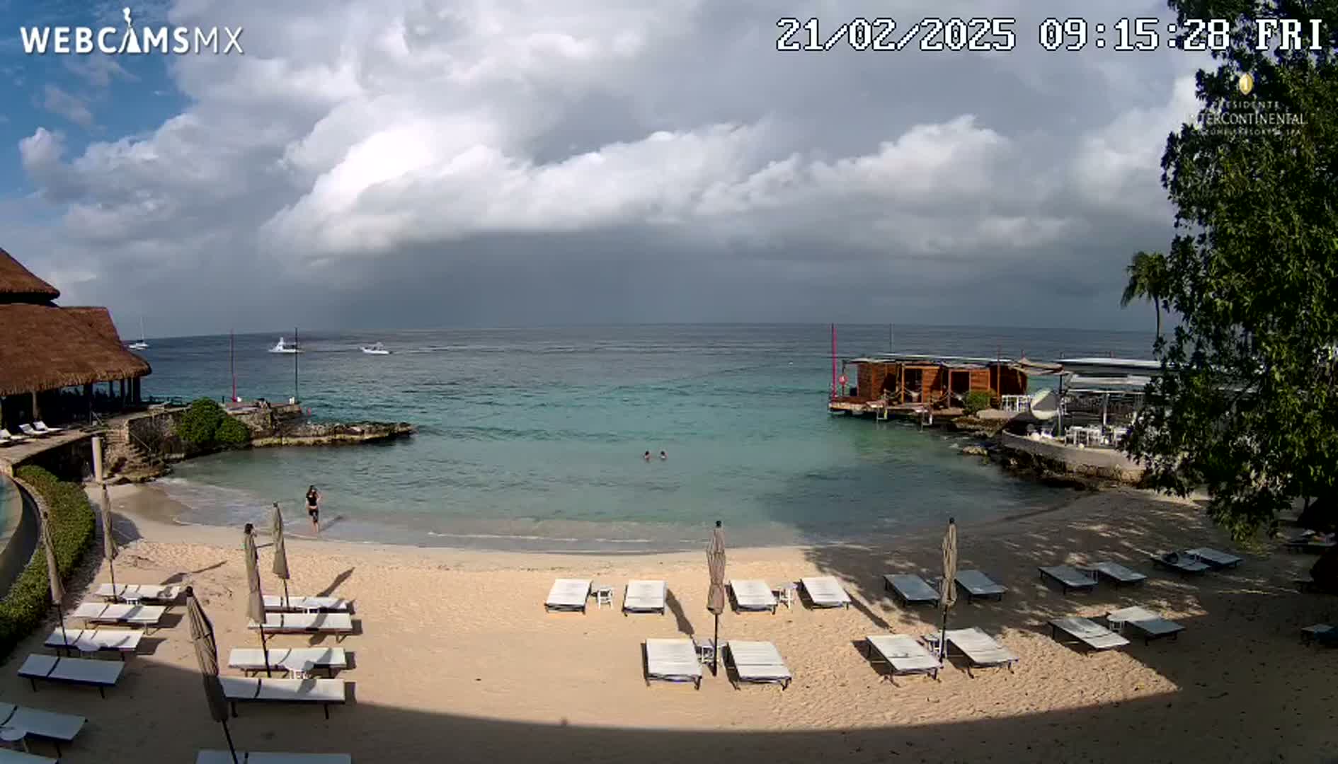 A partly cloudy day at a beach resort with lounge chairs on a sandy beach, calm turquoise water, and several small boats visible in the distance.