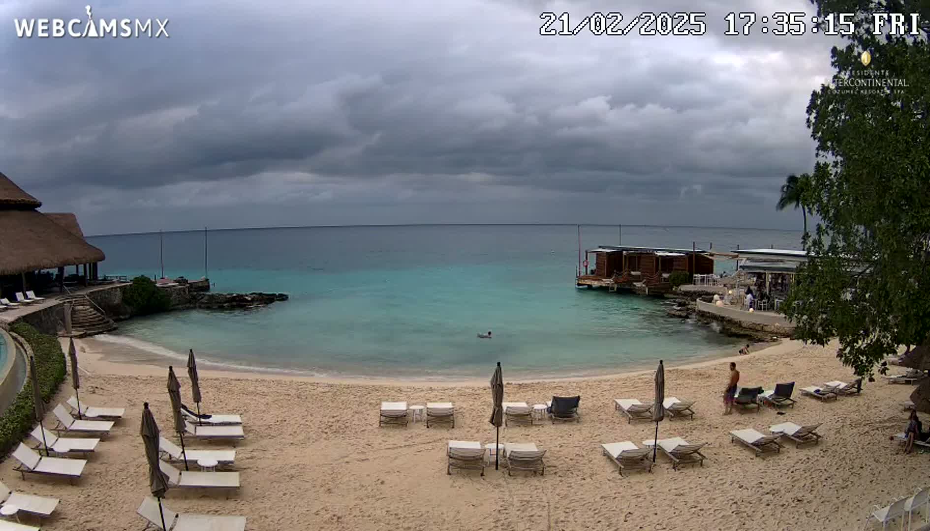 A sandy beach with lounge chairs and umbrellas under an overcast sky overlooks calm, turquoise water; a small overwater structure is visible in the distance.