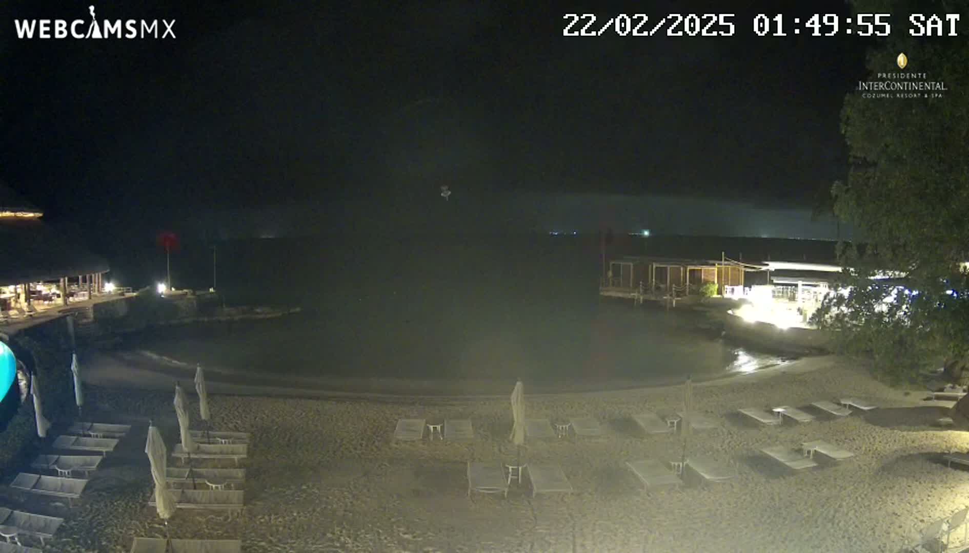 A nighttime beach scene shows a calm body of water, a sandy shore with empty lounge chairs, and low-lit buildings in the background.