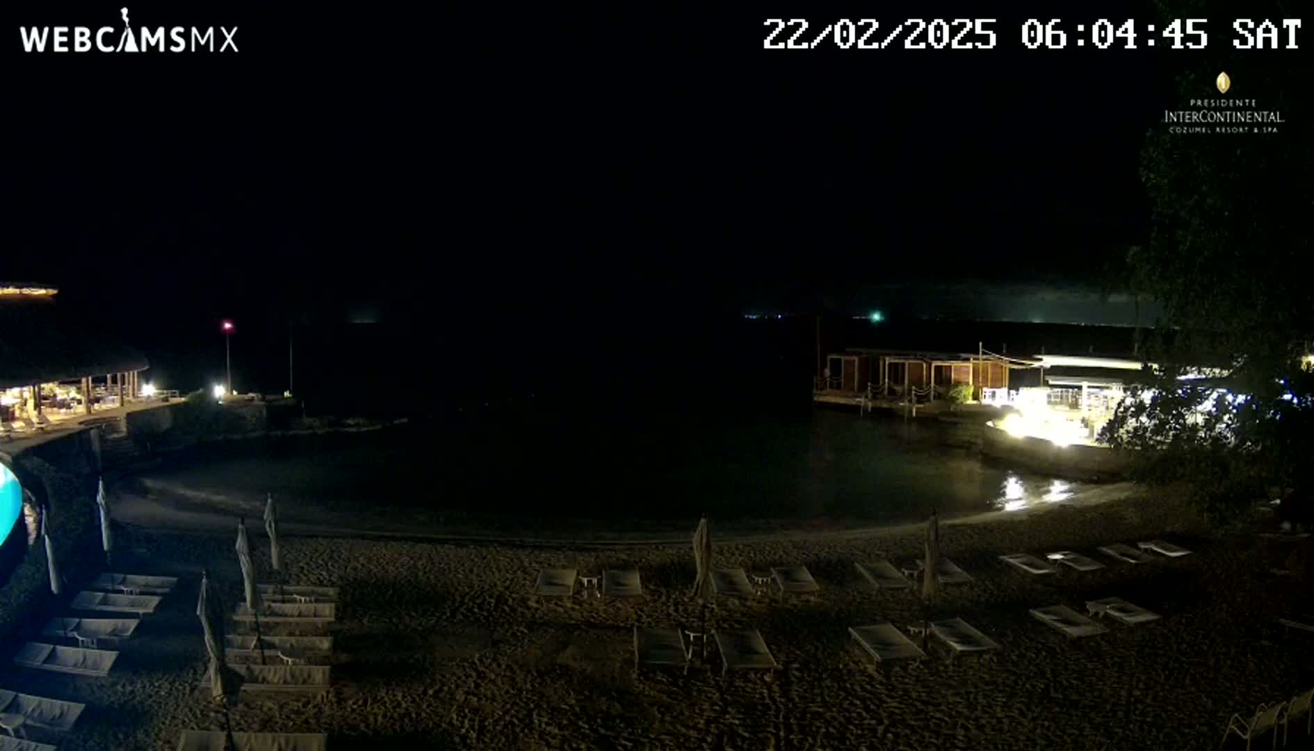 A nighttime beach scene shows a dark sandy beach with rows of empty lounge chairs, calm water, and low-lit buildings under a clear, dark sky.