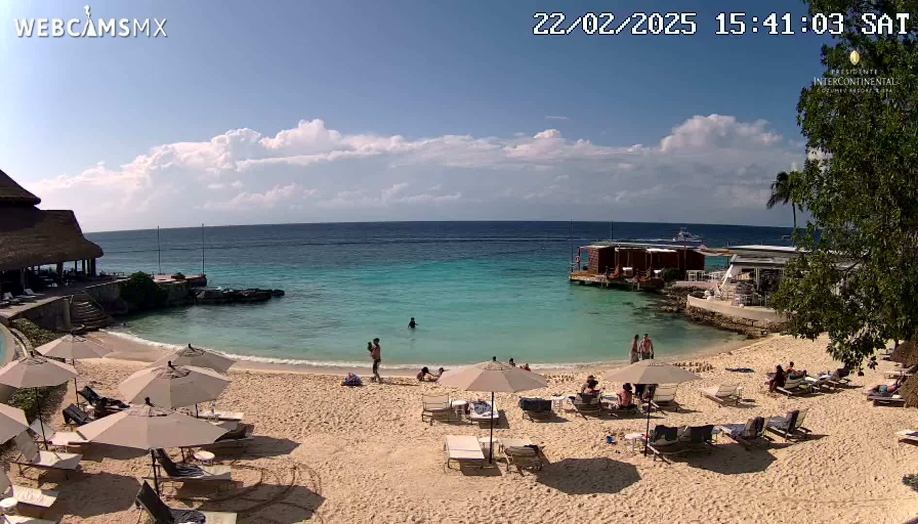 A sandy beach with turquoise water, beach umbrellas, lounge chairs, and several people relaxing under mostly sunny skies.