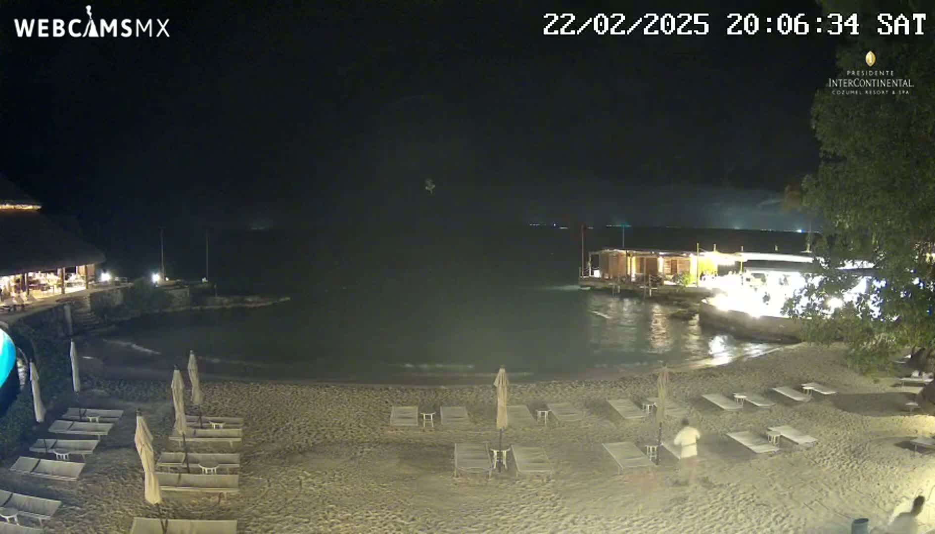 A nighttime beach scene with calm water, sand, lounge chairs under umbrellas, and buildings with lights in the background.