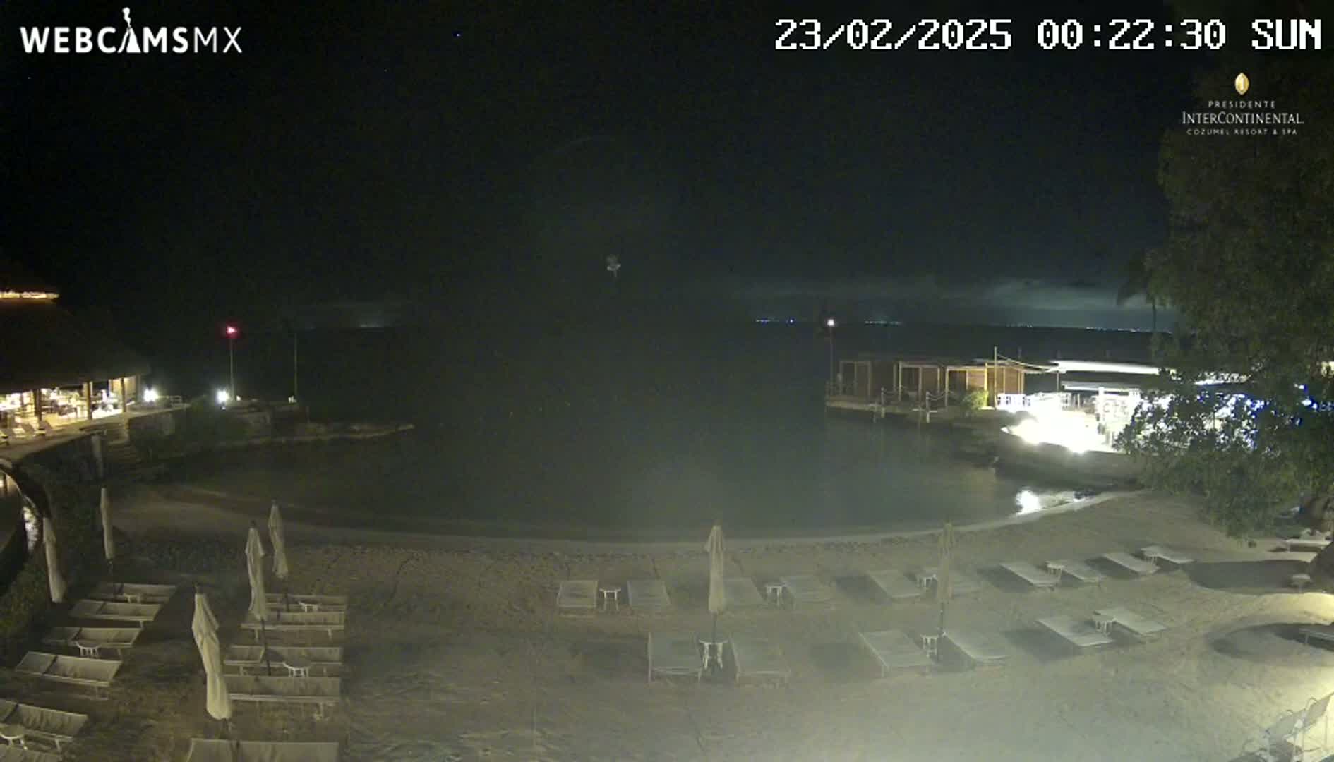 A nighttime beach scene shows a calm body of water, empty lounge chairs on a sandy beach, and low-level lighting around buildings under a mostly clear sky.