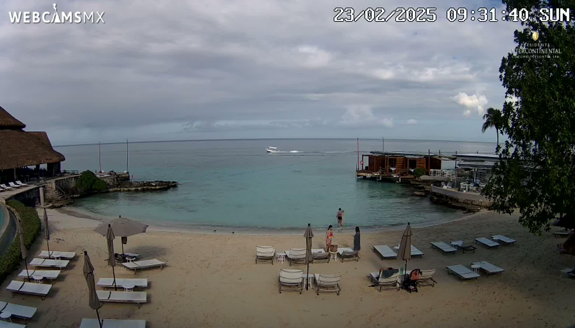 A sandy beach with lounge chairs and umbrellas, a small calm body of water, a wooden overwater structure, and a few people are visible on an overcast day.