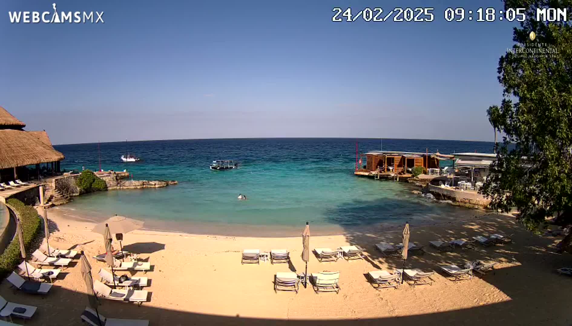A sandy beach with lounge chairs and umbrellas sits next to calm, clear turquoise water, under a sunny blue sky; a few boats are visible on the water.