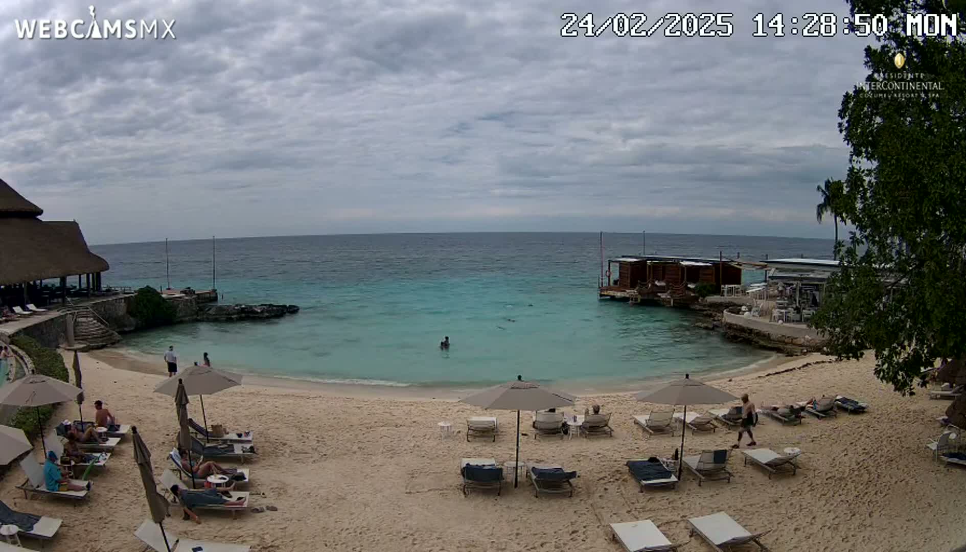 A sandy beach with lounge chairs and umbrellas under an overcast sky overlooks a calm turquoise ocean with a small overwater structure.