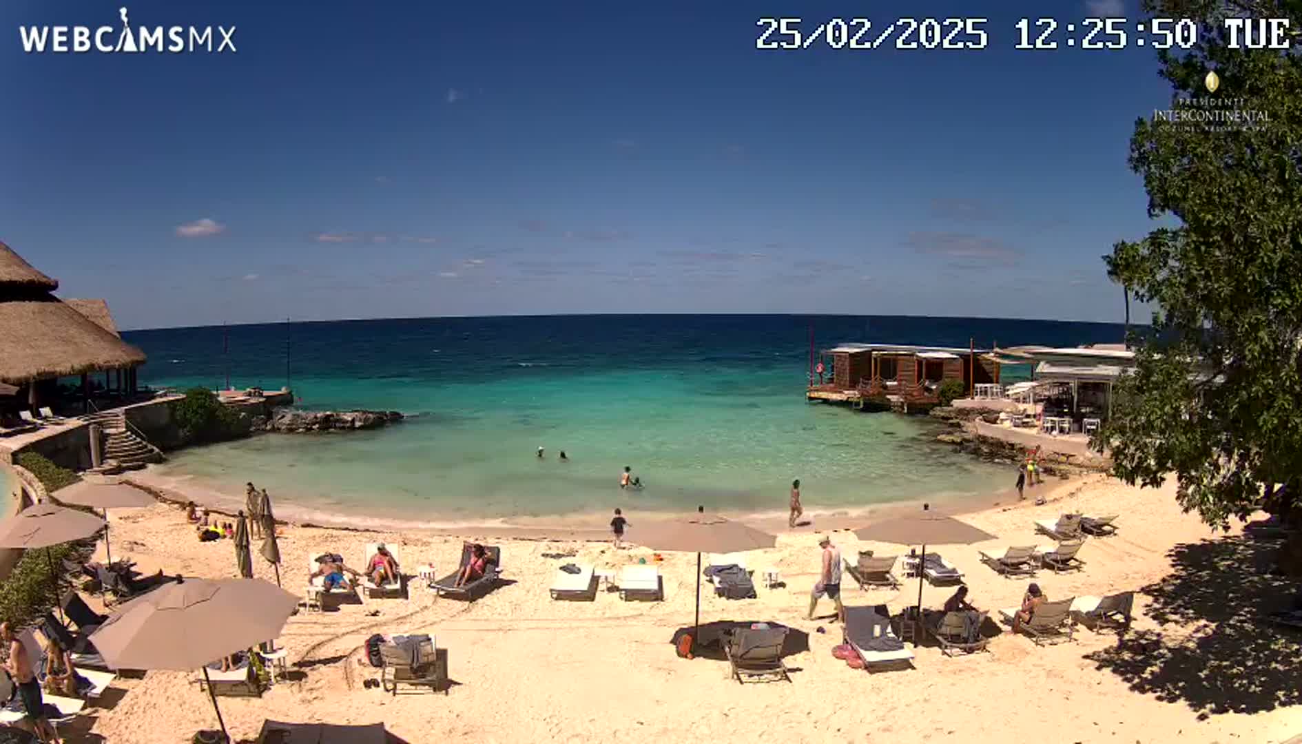 A sandy beach with lounge chairs and umbrellas, a calm turquoise ocean, and a few people swimming and sunbathing under a clear blue sky.