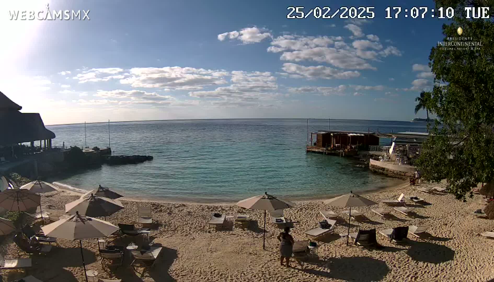 A sandy beach with lounge chairs and umbrellas under a partly cloudy blue sky overlooks calm turquoise water.