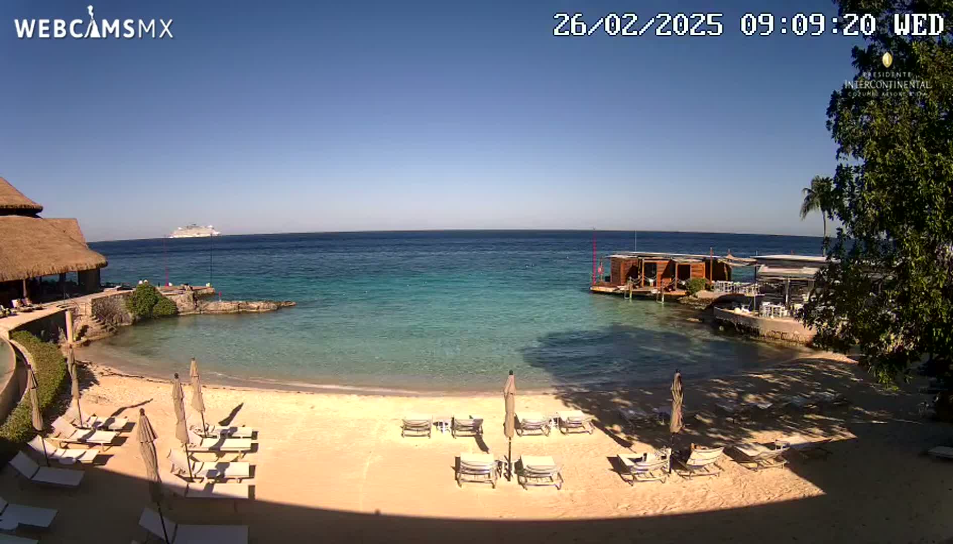 A sandy beach with lounge chairs and umbrellas sits beside a calm, clear turquoise ocean under a sunny sky, with a large cruise ship visible in the distance.
