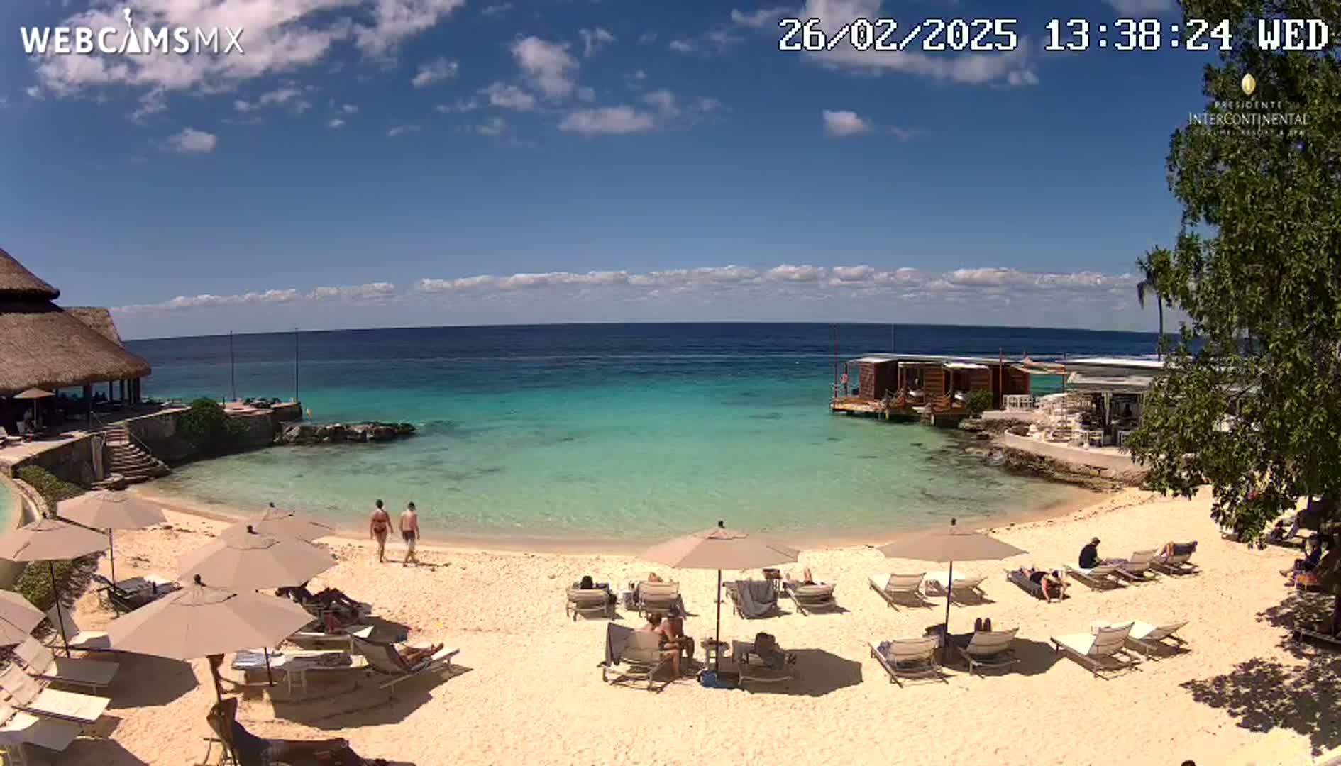 A sunny beach scene with turquoise water, a sandy shore lined with lounge chairs and umbrellas, and a few people relaxing.