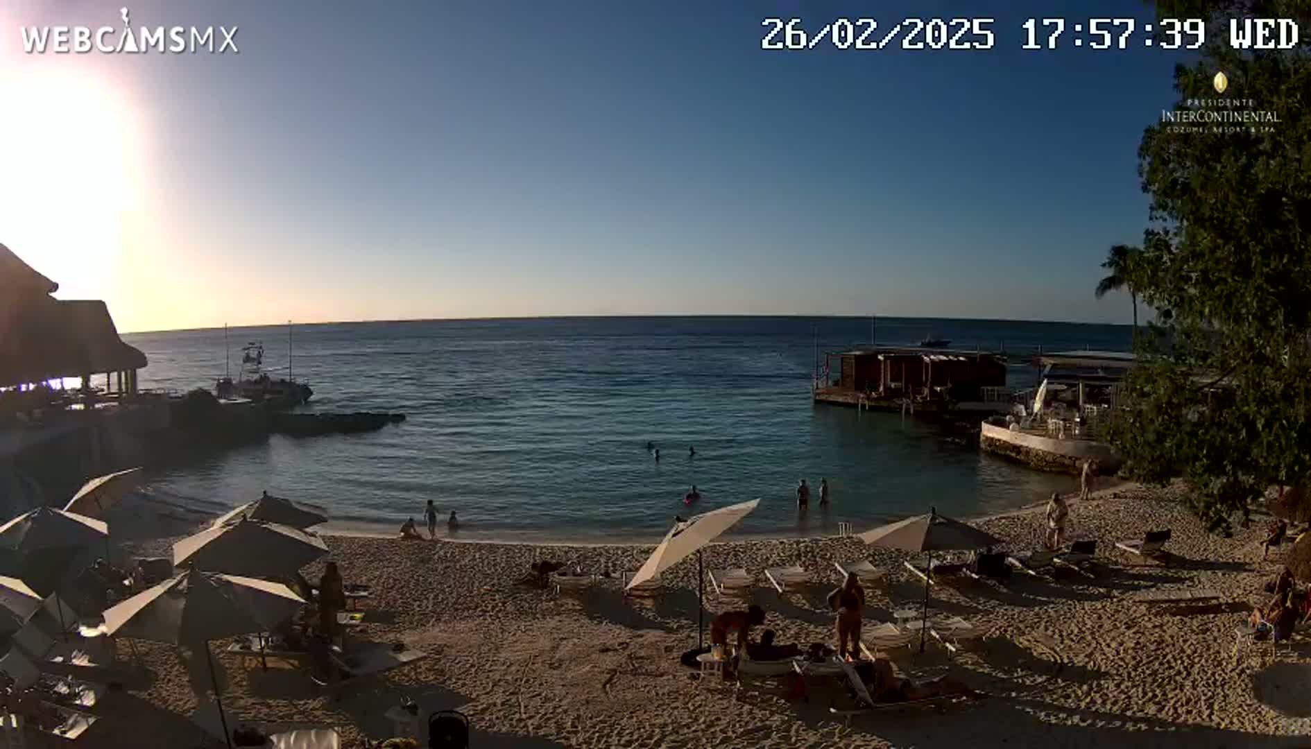A sandy beach with calm, clear water at sunset, showing people relaxing on lounge chairs under umbrellas near a calm ocean.