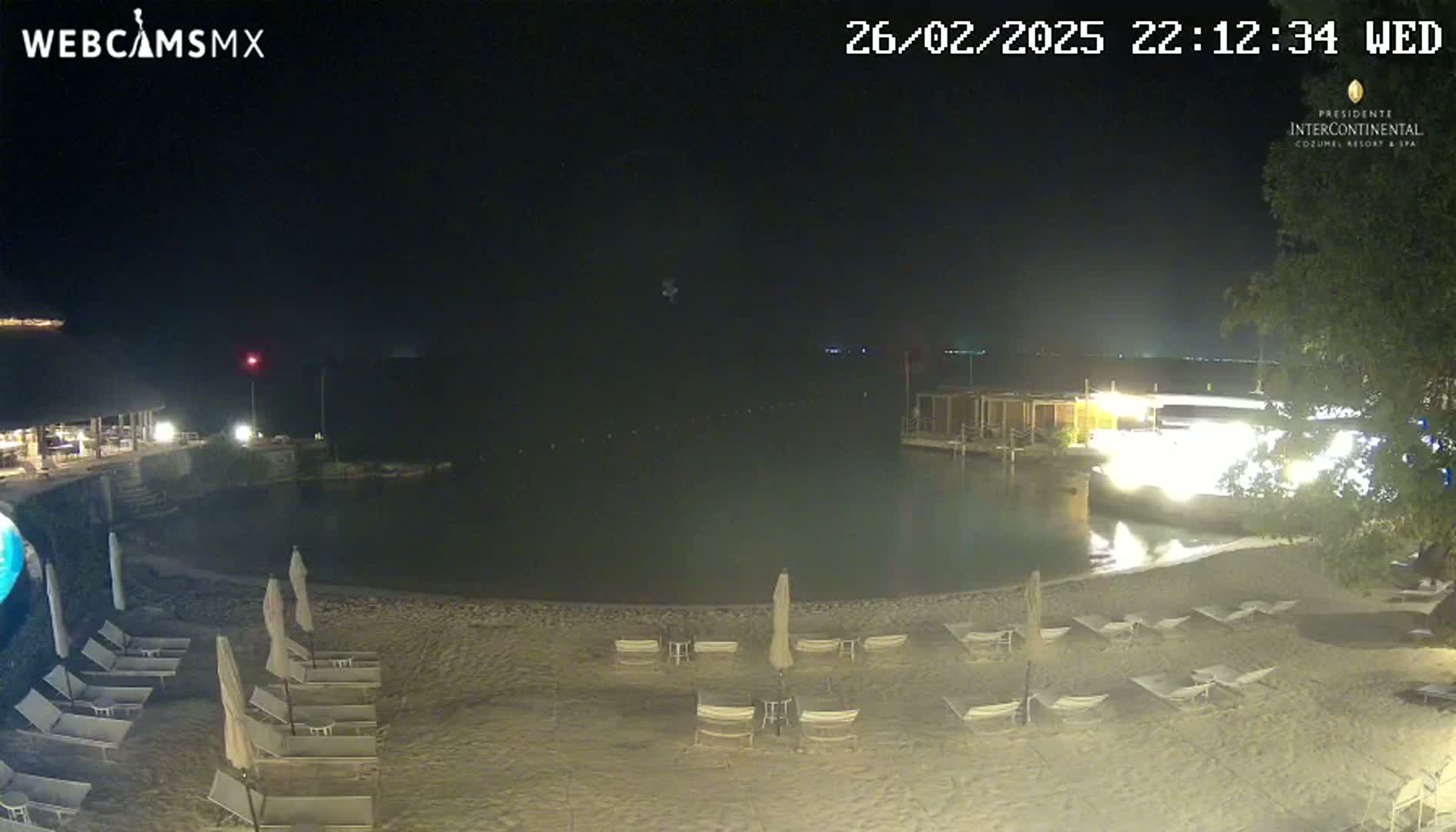 A nighttime beach scene shows a calm body of water, a sandy shore with lounge chairs and umbrellas, and a building with lights illuminated in the background.