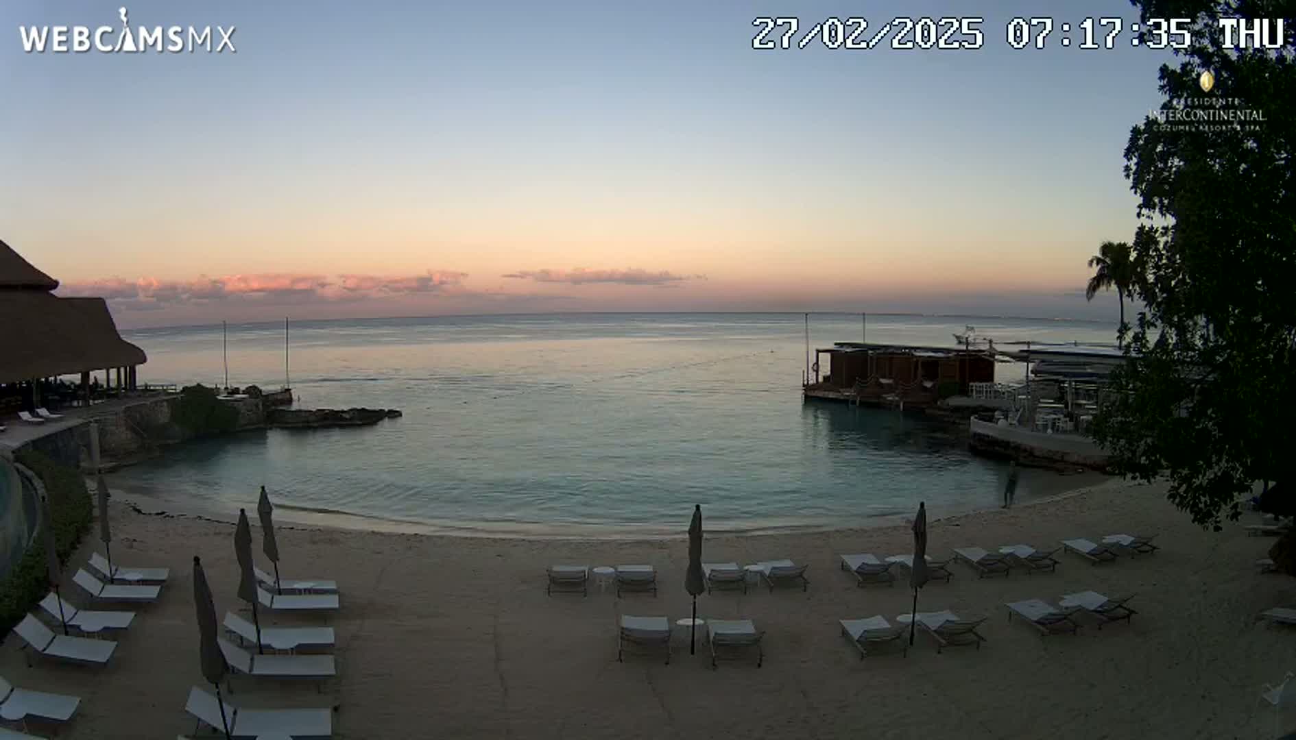 A tranquil beach scene at sunset shows calm ocean water, a sandy shore lined with lounge chairs and umbrellas, and a small building on the water.