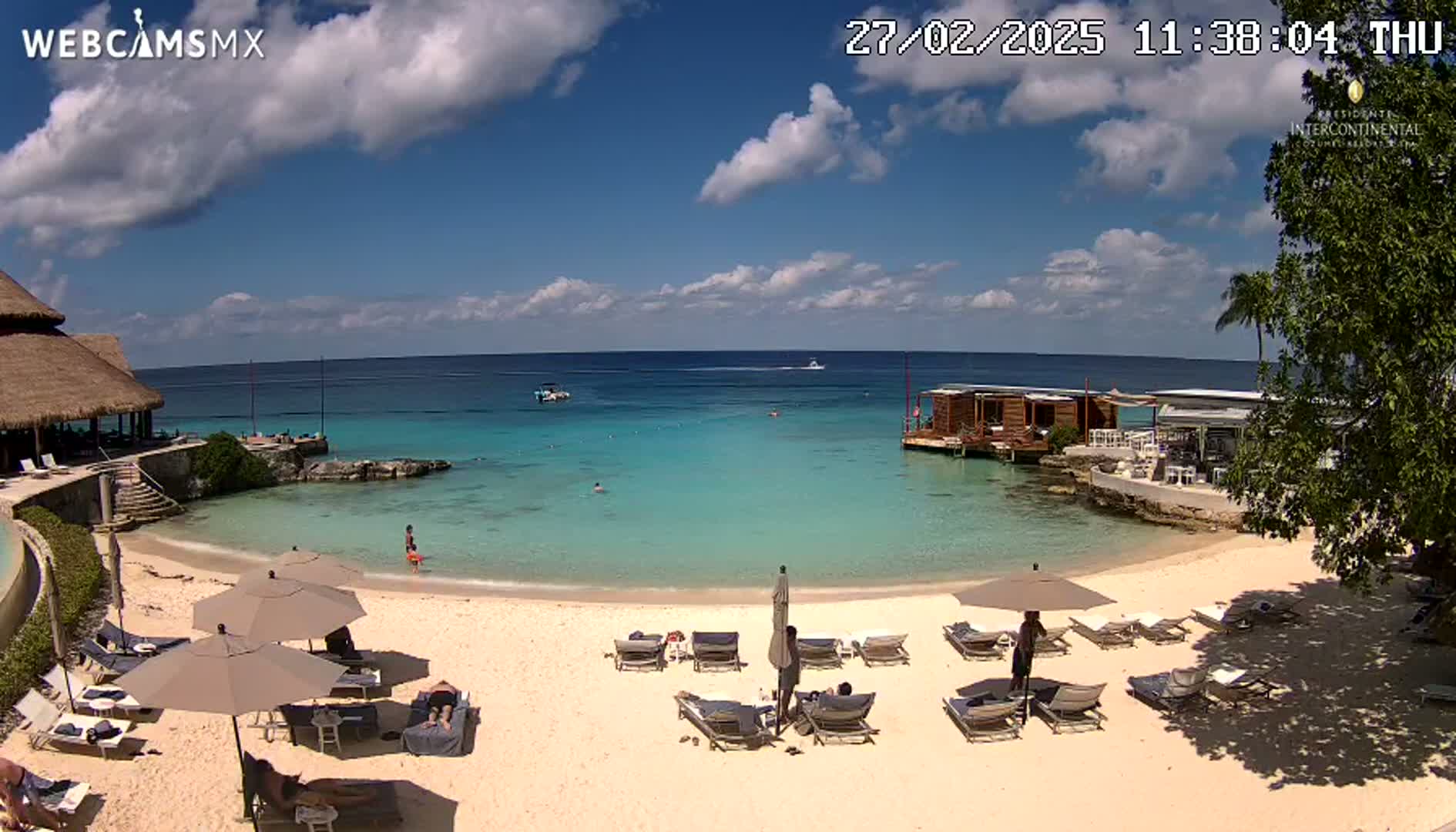A partly sunny day at a beach with turquoise water, showing lounge chairs under umbrellas, a small building on the water, and a thatch-roof structure on the shore.