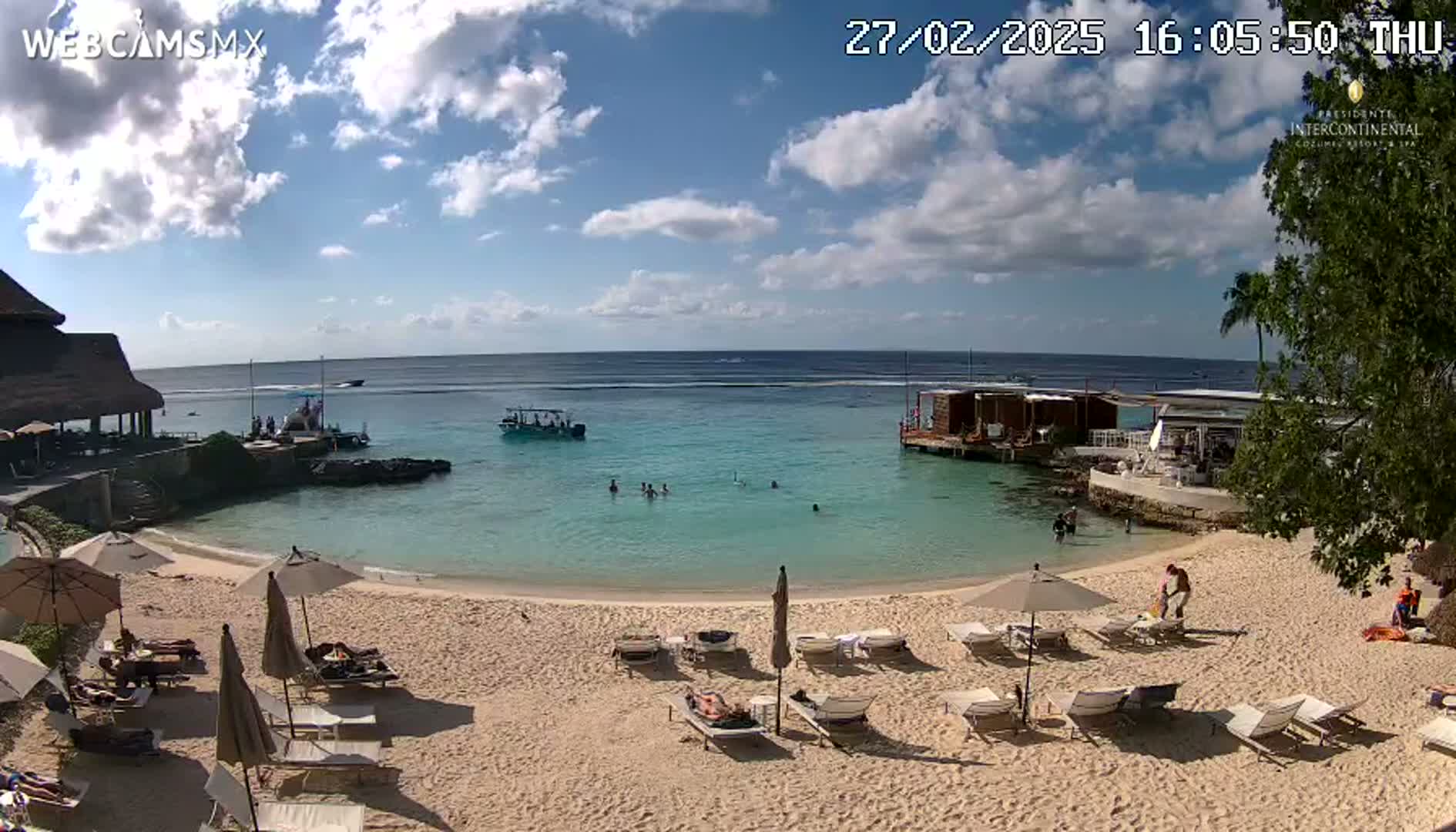 A sandy beach with lounge chairs and umbrellas under a partly cloudy sky overlooks calm, turquoise water with several small boats and people swimming.