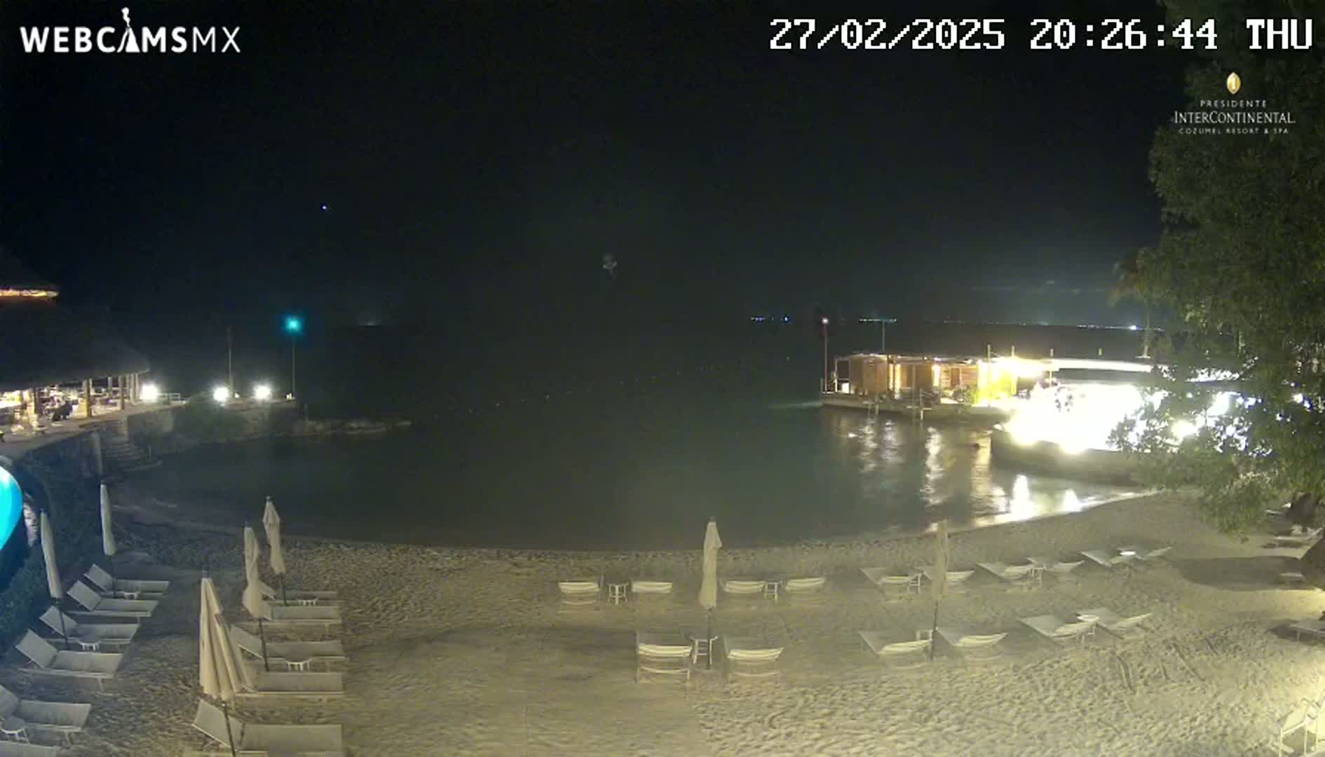 A sandy beach at night with several lounge chairs and umbrellas, illuminated buildings along the water's edge, and calm water under a clear, dark sky.