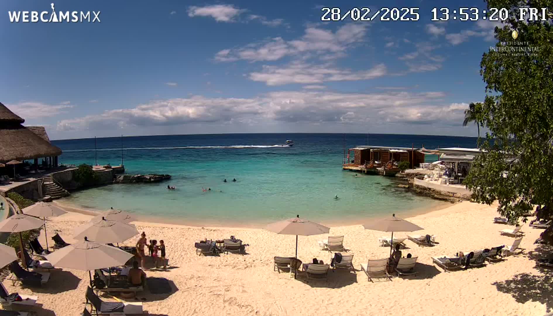 A sunny day at a beach resort shows a sandy beach with lounge chairs and umbrellas, a calm turquoise ocean with a few people swimming, and some buildings on the shore.