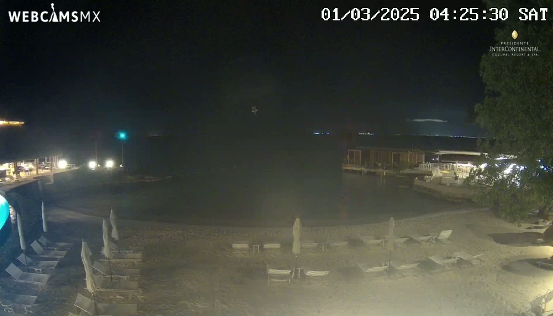 A nighttime beach scene shows a calm body of water, a sandy shore with empty lounge chairs and umbrellas, and some low-lying buildings in the background.