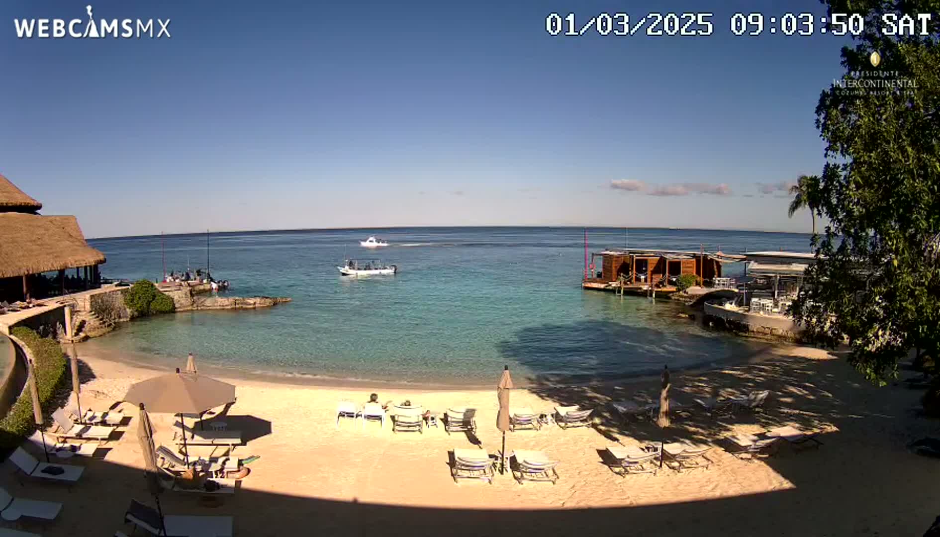 A sandy beach with lounge chairs and umbrellas overlooks calm, clear turquoise water, with boats in the distance under a bright, sunny sky.