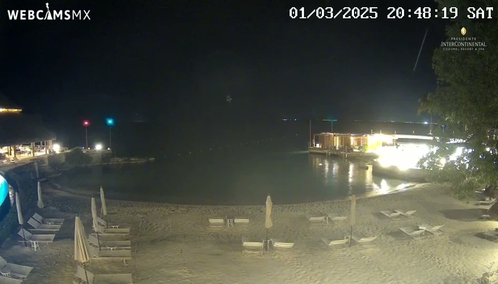 A nighttime beach scene shows a sandy shore with lounge chairs and umbrellas, a calm body of water, and buildings with lights illuminated along the edge of the water.