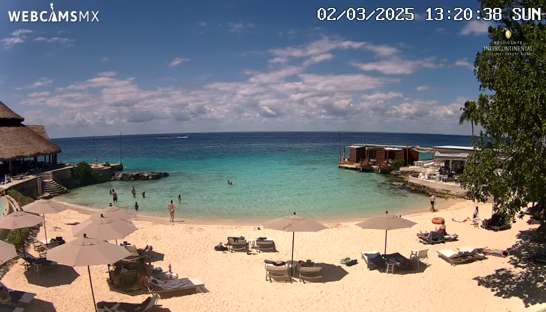 A sandy beach with turquoise water, several beach umbrellas, lounge chairs, and people swimming and relaxing under a partly cloudy sunny sky.