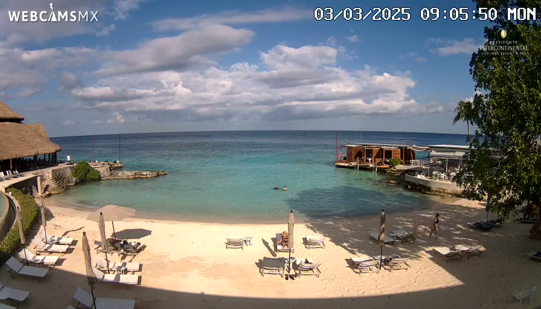 A partly cloudy day shows a beach with lounge chairs and umbrellas, calm turquoise water, and a building with a thatched roof.