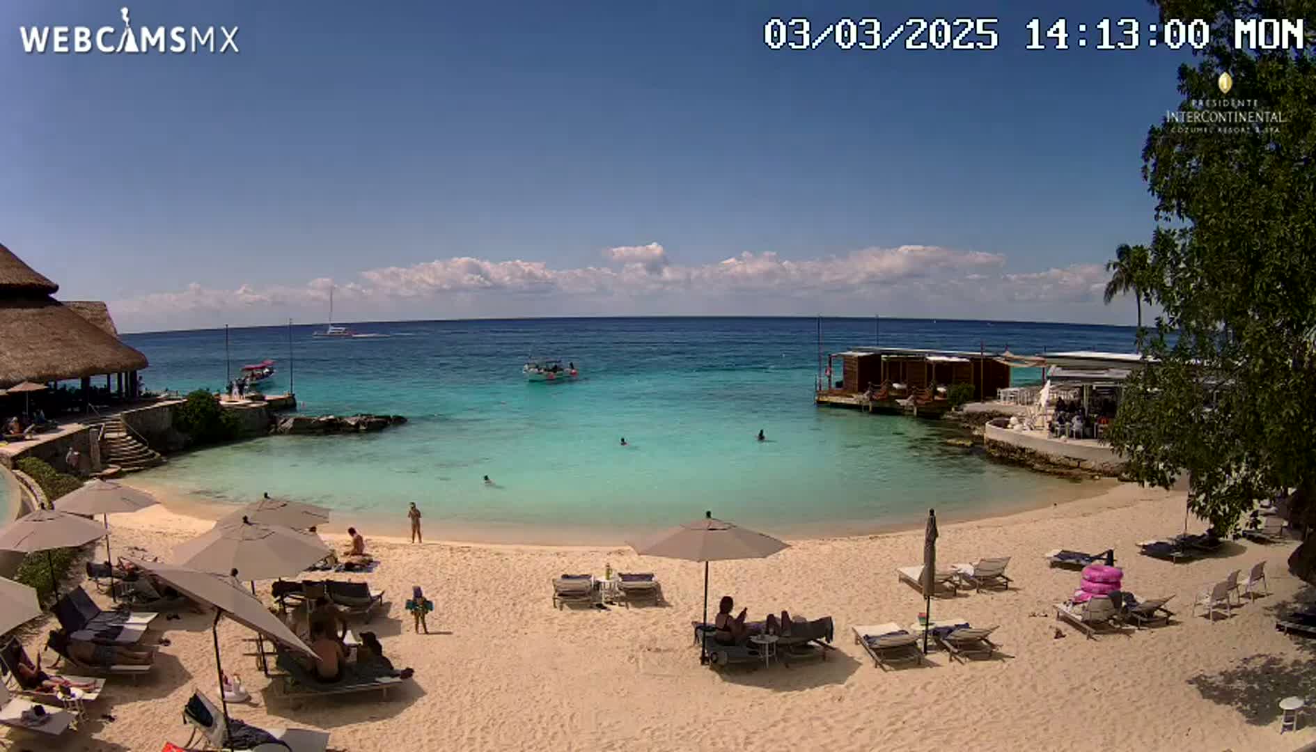 A sandy beach with lounge chairs and umbrellas under a mostly sunny sky, overlooks a calm, turquoise ocean with several small boats and people swimming.