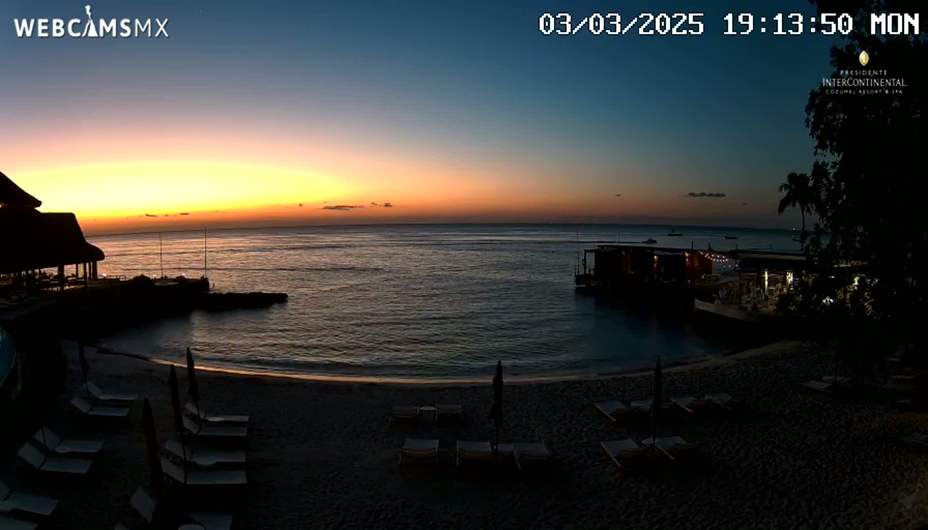 A beach at sunset with lounge chairs and a calm ocean, under a mostly clear sky.