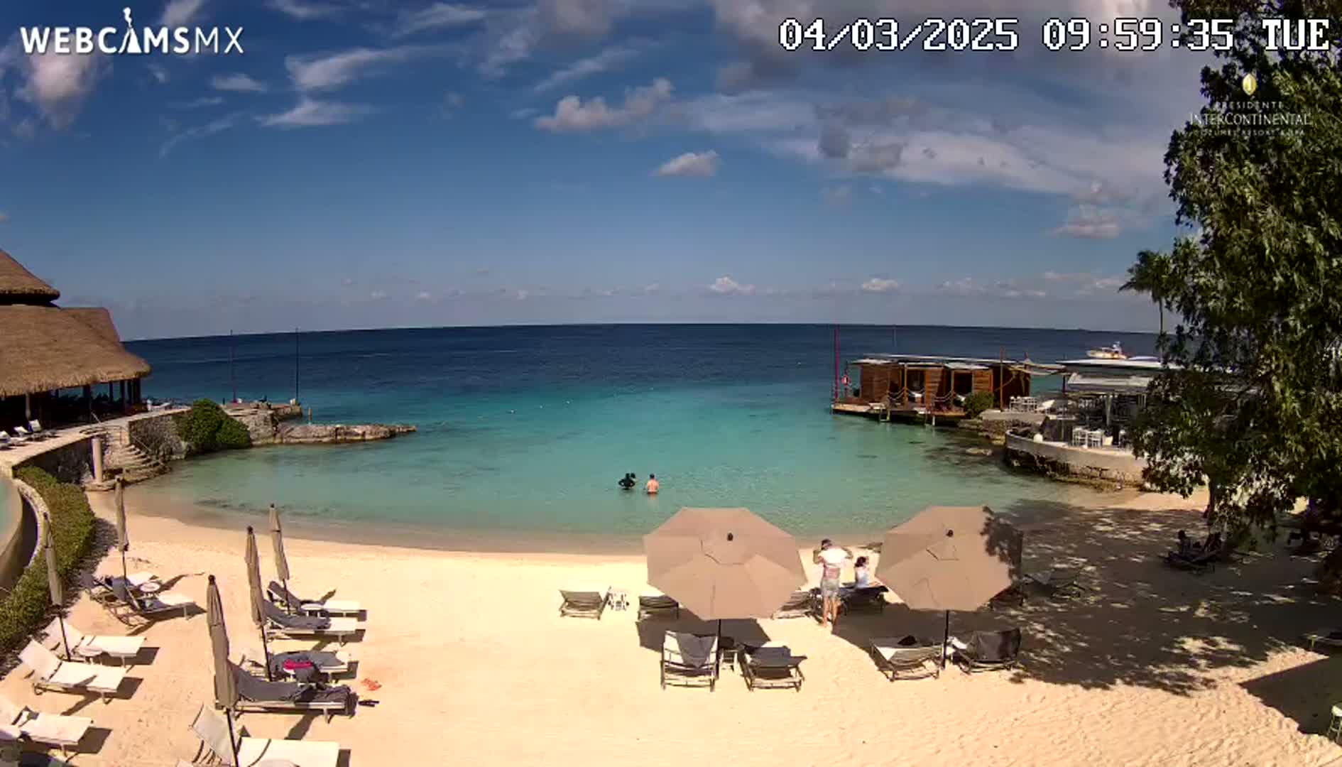 A sandy beach with lounge chairs and umbrellas is situated next to a calm, turquoise ocean under a partly sunny sky.