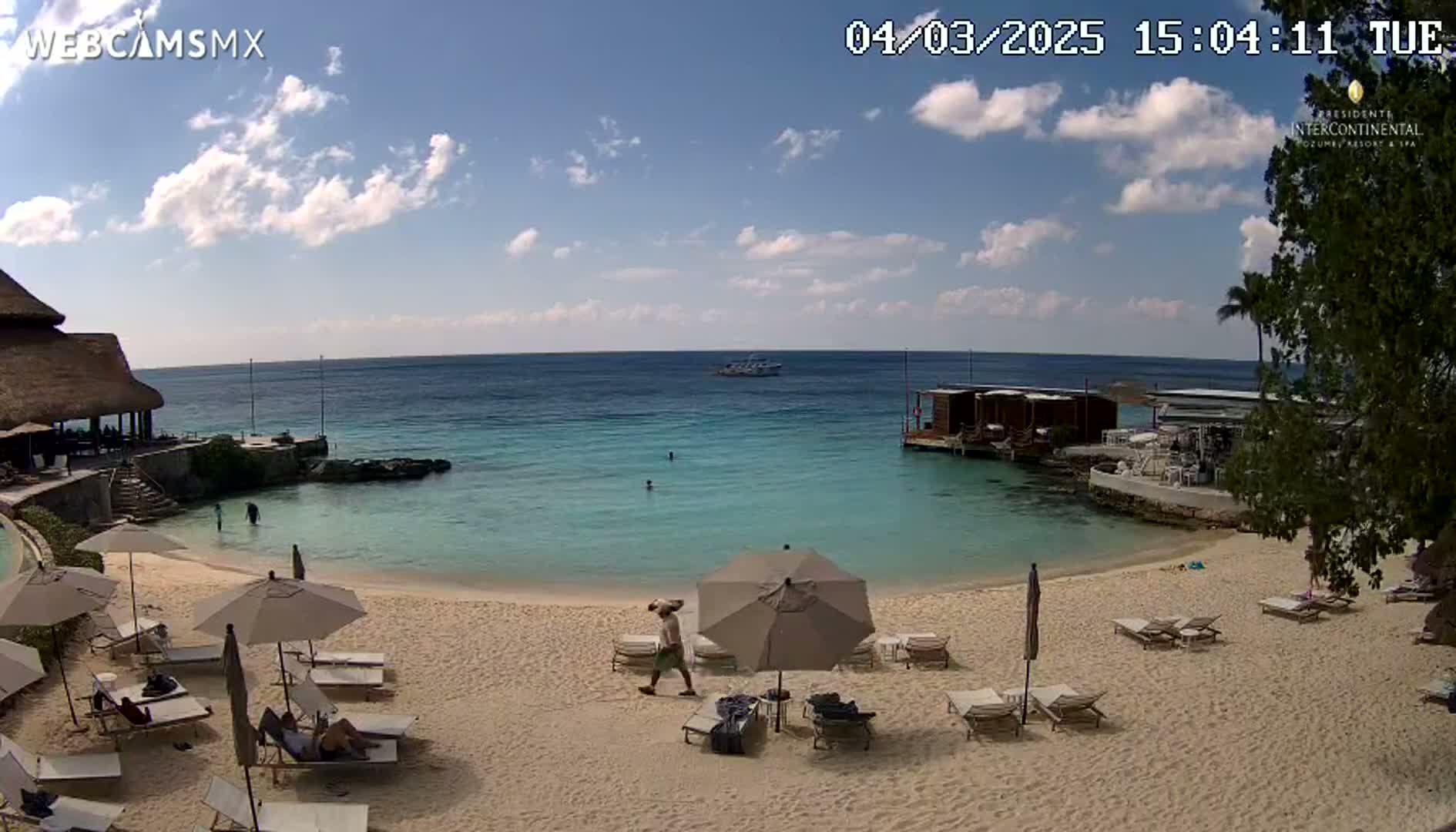 A sandy beach with lounge chairs and umbrellas under a partly cloudy sky, overlooks a calm turquoise ocean with a few people swimming and a boat in the distance.