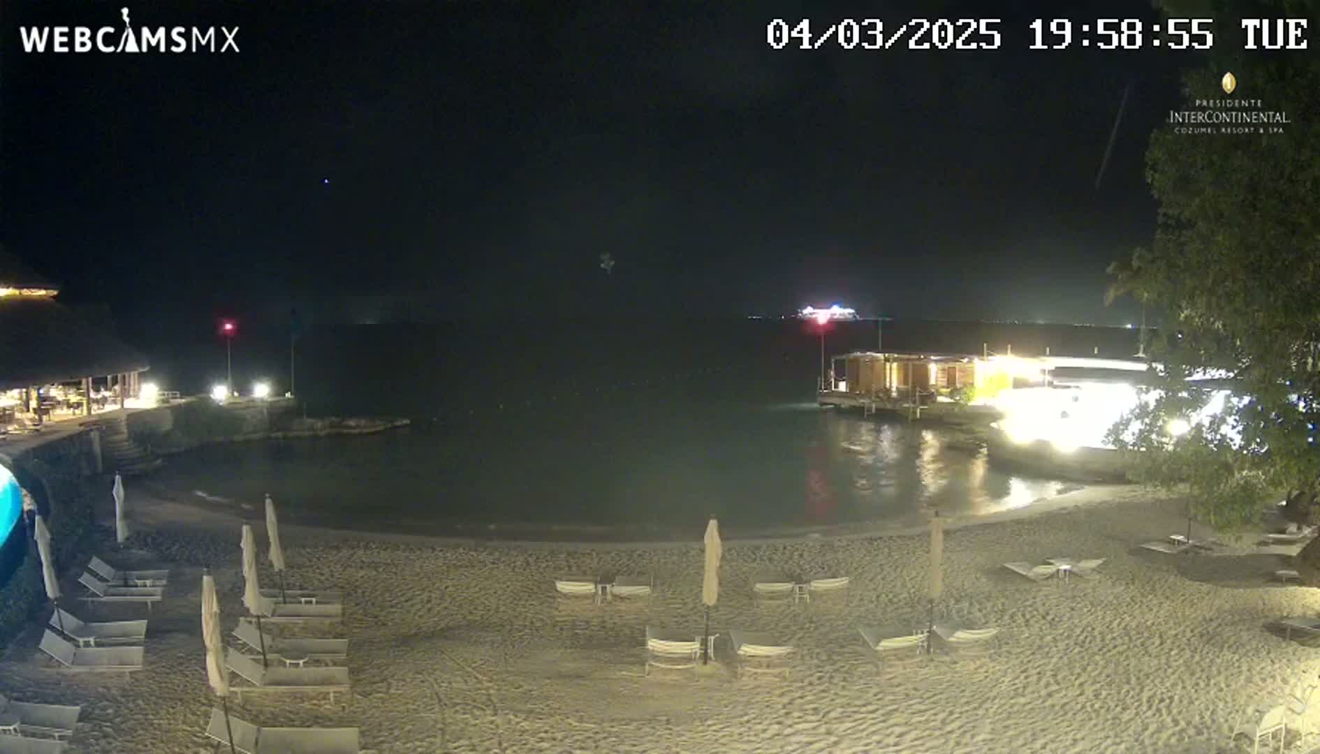 A nighttime beach scene shows a sandy shoreline with lounge chairs and umbrellas, a calm body of water, and buildings with lights illuminated in the background under a clear, dark sky.