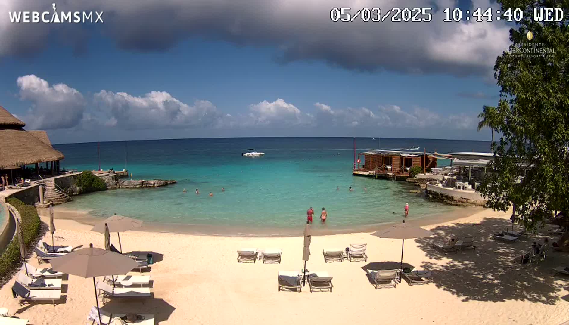 A partly sunny beach scene shows a sandy shore with lounge chairs and umbrellas, a calm turquoise ocean with a few people swimming, and a thatched-roof building on the left.