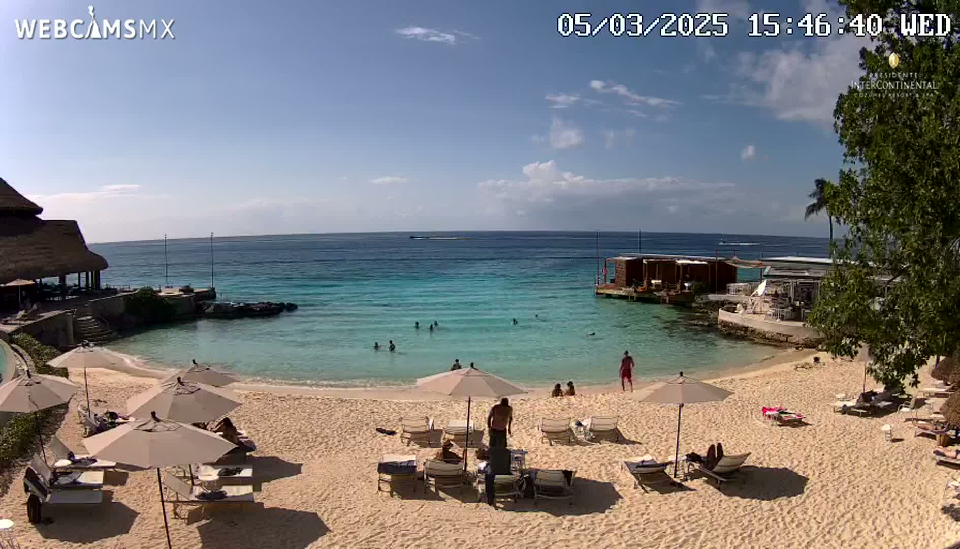 A sandy beach with turquoise water, lounge chairs under umbrellas, and several people swimming and relaxing on a sunny day.