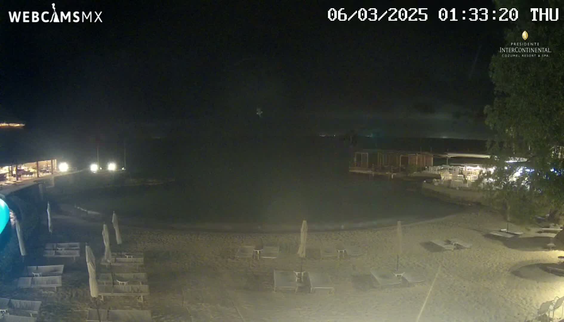 A nighttime view of a resort beach with a dark pool, lounge chairs, and umbrellas under a cloudy sky.