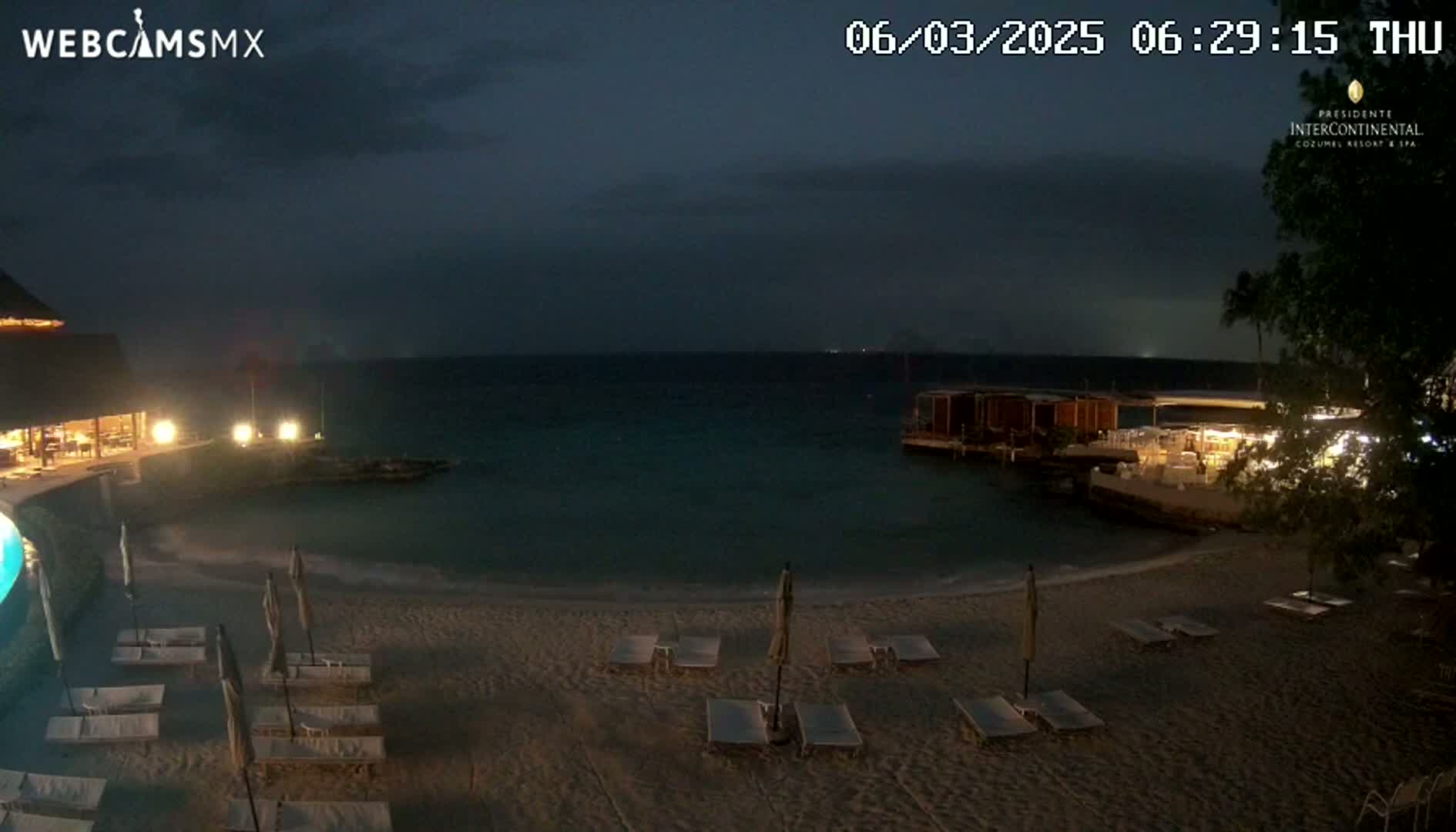A dark, nighttime beach scene shows a sandy shore with empty lounge chairs and umbrellas under an overcast sky, overlooking a calm body of water with a restaurant visible in the distance.