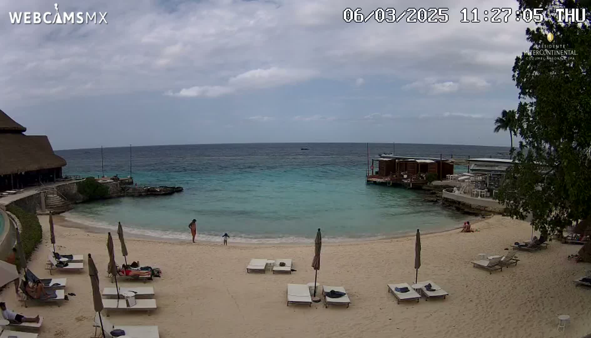 A partly cloudy day at a beach with turquoise water, showing lounge chairs, umbrellas, and a few people on the sand and in the water near a beachfront building.